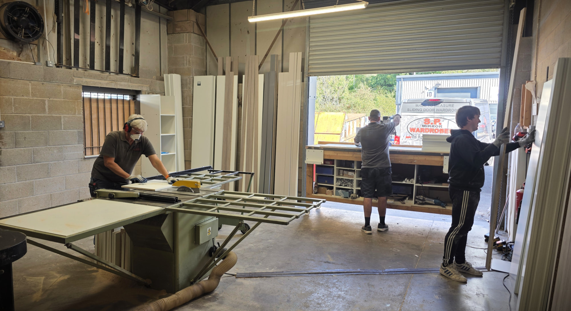 Three of the team at work in the SP Mirror Wardrobes workshop in Pontypridd, with boards stacked by the benches and the company van outside