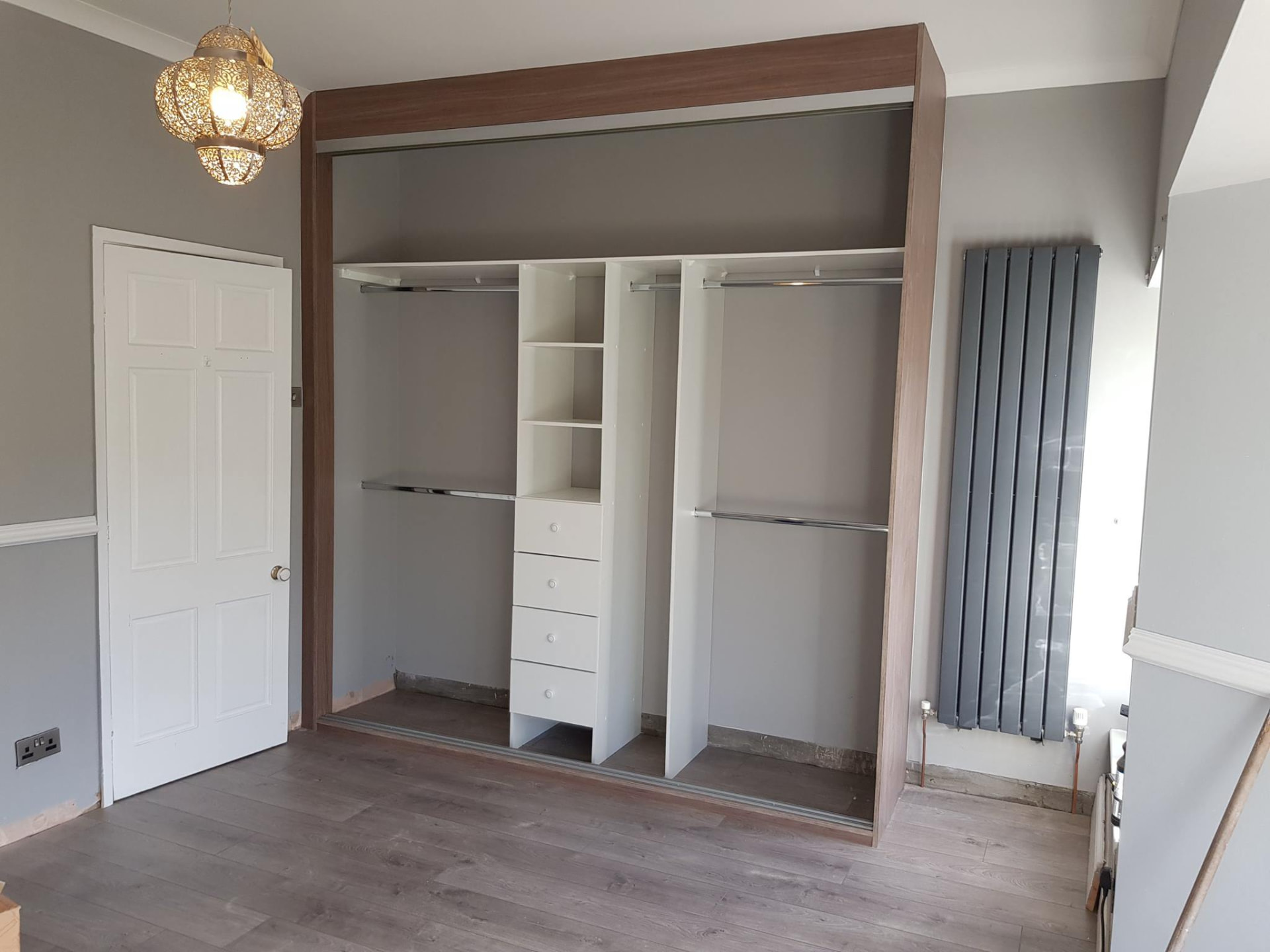 Open white wardrobe interior with double hanging on both sides, central shelves and drawers, and an oak-effect surround before the sliding doors are fitted in Carmarthen